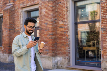 Young man walking in the city reading messages on the phone smiling, with a cup of hot drink, businessman entrepreneur programmer in casual clothes using an application on a smartphone