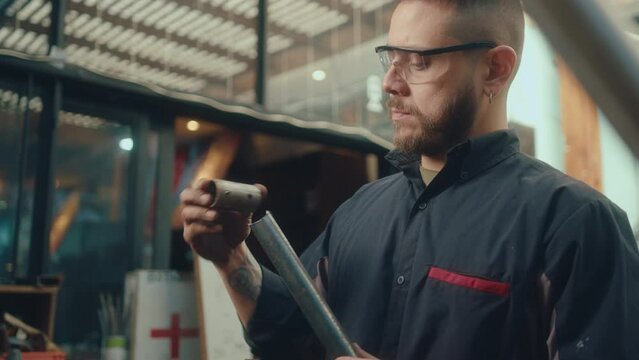Young repair shop worker in safety glasses and workwear connecting metal detail and pipe, then fastening workpiece with workbench vise and taking angle grinder. Tilt-down shot
