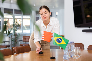 Positive young woman putting little flag of Bahrain on the table with flag of Brazil in conference...