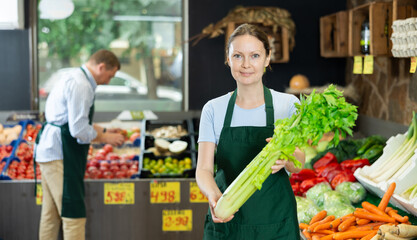 Young positive female seller in apron displaying assortment of celery at supermarket