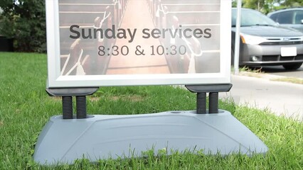 sunday services 830 and 1030 time sign information on grass lawn next to sidewalk and street road in summer on bright day with car vehicle pulling up and parking and traffic and person passing