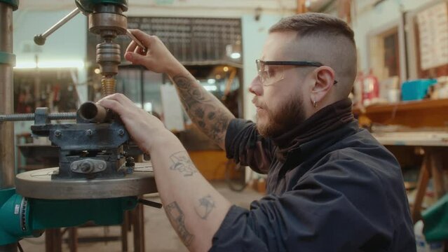 Young bearded metalworker in safety glasses using drill press machine on metal pipe at work in repair garage