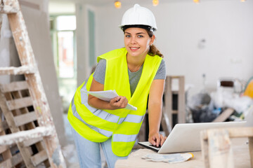Female foreman in a protective helmet and a yellow vest checks the execution of repair work using a laptop