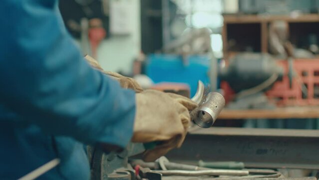 Close up shot of metalsmith in protective gloves using hammer and wire brush on metal workpiece after welding in the workshop