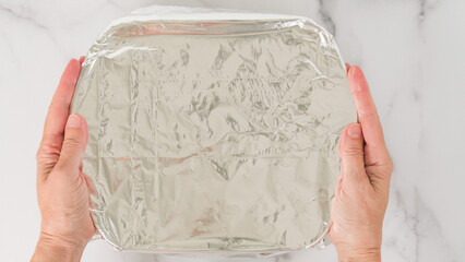 The chef hands holding a baking dish covered with aluminum foil. Close-up view from above