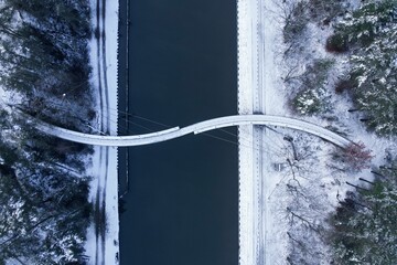 Top view of a curving bridge over the calm lake connecting the two edges of the snowy forest