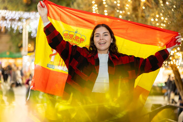 Happy young female tourist visiting traditional festive street Christmas fair in Barcelona, posing with national Spanish flag in hands on background of lighted stalls - obrazy, fototapety, plakaty