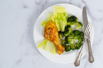 A plate of an oven-baked chicken drumstick and broccoli, served with fresh lettuce, close-up on a marble kitchen table, flat lay