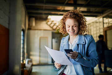 Young woman looking at mobile phone and holding document in office