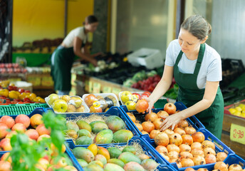 Positive skilled saleswoman in green apron working in fruit and vegetable section of supermarket,...