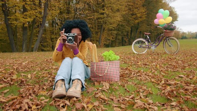 Smiling cute woman with camera in autumn park, sitting on lawn taking picture of photographer. Shopping bag with products and decorated bike on background. High quality 4k footage