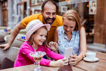 Little girl holding cell phone with parents in outdoor cafe