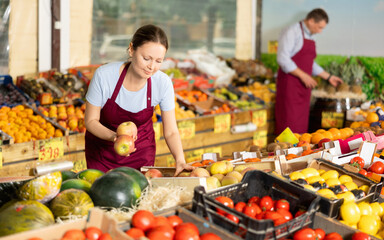 Obraz premium Interested positive saleswoman arranging sweet juicy organic apples on fruit and vegetable stall in greengrocery..