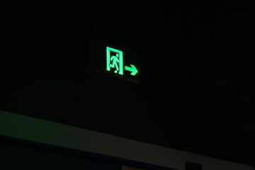 Luminescent green emergency exit sign with arrow in the dark - obrazy, fototapety, plakaty