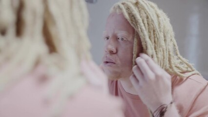 Reflection of an albino man with dreadlock hair looking at himself in the mirror with his hand on his face. Cinematic 4k.