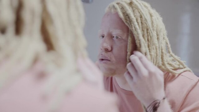Reflection of an albino man with dreadlock hair looking at himself in the mirror with his hand on his face. Cinematic 4k.