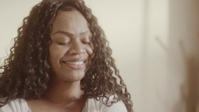 Linda mulher de cabelos cacheados, sorrindo, sentada na cama vestindo pijama. Cinematico 4k.