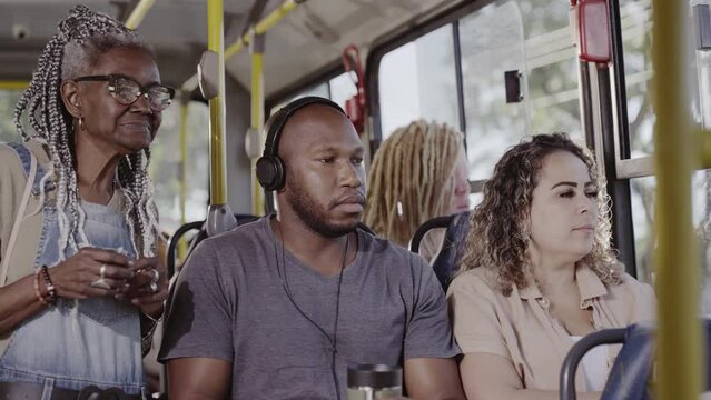 Young black man wearing headphones, standing up on a crowded bus to give up his seat to an elderly woman. Cinematic 4k.