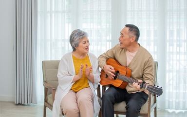 Asian sweet senior elderly couple wearing casual clothes, staying in indoor cozy home at living room, playing guitar, singing together with happiness, smiling. Retirement, Healthcare Concept.