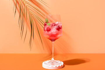 Decorative podium with glass of fresh raspberry mojito and palm leaf on orange background