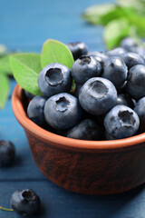 Tasty fresh blueberries with leaves in bowl on blue wooden table, closeup