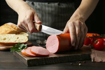 Woman cutting tasty boiled sausage at dark wooden table, closeup - obrazy, fototapety, plakaty