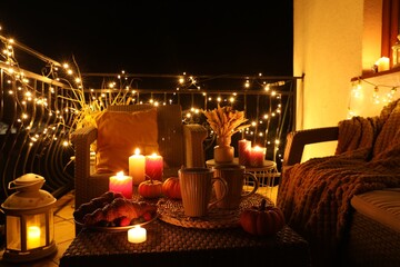 Rattan furniture, cups, fairy lights, burning candles and other autumn decor on outdoor terrace at...
