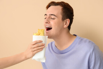 Young man eating tasty sandwich wrap on beige background, closeup