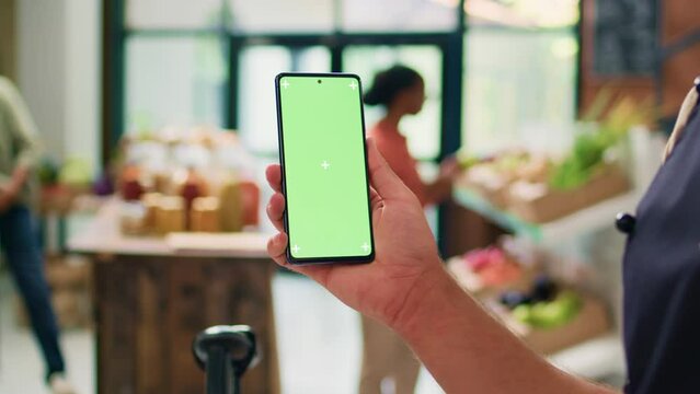 Owner uses greenscreen on smartphone, showing isolated chromakey template on device in local organic zero waste eco market. Young vendor presents copyspace mockup display.