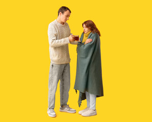 Young man giving cup of hot tea to his ill girlfriend on yellow background