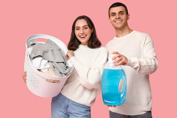 Young couple with laundry basket and bottle of detergent on pink background
