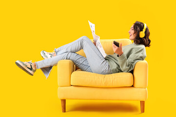 Young woman with newspaper and mobile phone in armchair on yellow background - obrazy, fototapety, plakaty