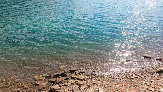 the sparkle of day sunlight reflected on the blue water's surface on a peaceful sunny day. slow water ripple on rocky shore. beautiful pure water full of minerals. natural landscape footage background