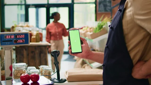Young man holds smartphone at store checkout, presenting isolated greenscreen display on mobile device. Farmer and vendor using blank chromakey template with copyspace technology.