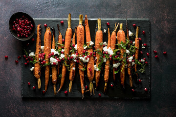 A black slate board of roasted carrots topped with pomegranate, herbs and cheese