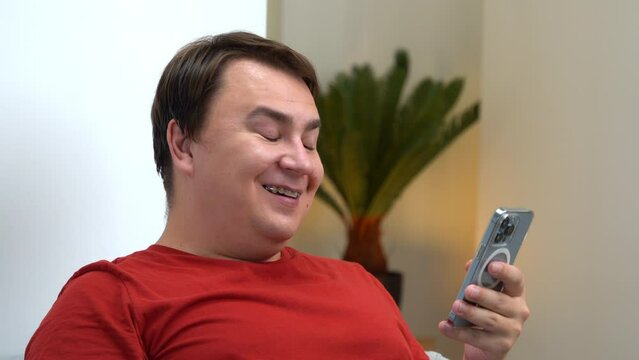 Portrait of man in red T-shirt sitting on sofa holding smartphone in hands, enthusiastically reading chat, smiling, laughing, distracted, waving head, hand gesture, grinning expression Armchair critic