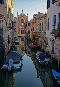 The still water of a canal in Venice, Italy reflects the facade of Madonna dell’Orto church. The painter Tintoretto is buried there. Buildings and boats in early morning in a scene of serenity.