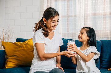 A loving Asian mother shares a glass of milk with her daughter on the sofa creating a heartwarming...