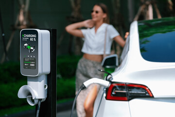 Young woman travel with EV electric car charging in green sustainable city outdoor garden in...