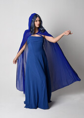Naklejka premium Full length portrait of beautiful female model wearing elegant fantasy blue ball gown and flowing cape with hood. Standing pose, with gestural arms reaching out . Isolated on white studio background.