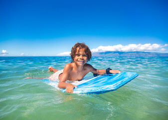 Smiling diverse young boy boarding in the beautiful blue ocean. Enjoying a fun day playing at the beach while on vacation