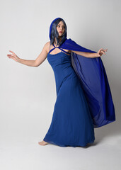 Full length portrait of beautiful female model wearing elegant fantasy blue ball gown and flowing cape with hood.
Standing pose, with gestural arms reaching out . Isolated on white studio background.