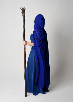Full length portrait of beautiful female model wearing elegant fantasy blue ball gown, flowing cape with hood.Standing pose walking away, holding a wooden wizard staff. Isolated on white studio backgr