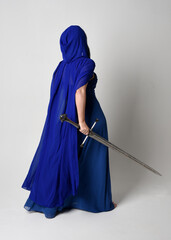 Full length portrait of beautiful female model wearing elegant fantasy blue ball gown, flowing cape with hood. Standing pose walking away, holding a sword weapon Isolated on white studio background. © faestock