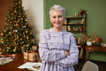Obraz na płótnie Canvas Smiling happy senior female interior designer in knitted lace cardigan standing with crossed hands, decorating house for christmas, standing next to table with lighted decorated tree on background