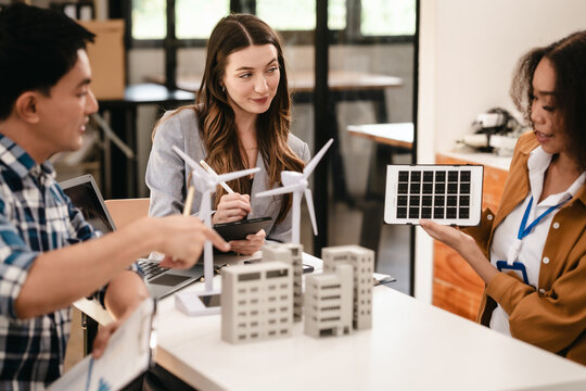 Diverse team with Asian man, African American woman, Caucasian woman discussing a clean energy city planning project with building models and solar panels.