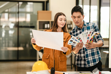 Asian man and Caucasian woman, both engineers, examining set of blueprints closely during team meeting, with house model on the table. Color swatches
