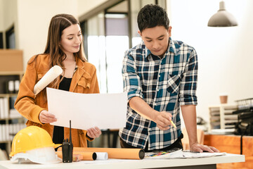 Asian man and Caucasian woman, both engineers, examining set of blueprints closely during team meeting, with house model on the table.