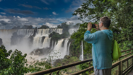 A man stands on the observation deck at the railing, taking pictures of the waterfall on his...