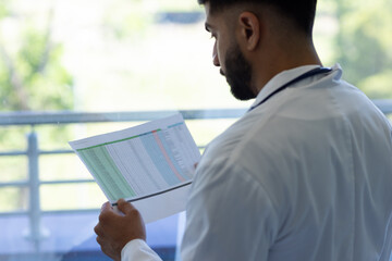 Biracial male doctor reading document in sunny hospital room